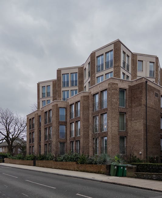 A multi-story residential building with a brick facade and large, vertically aligned windows in Hampstead. The building features a combination of traditional brickwork and modern architectural elements, with some sections displaying a lighter, beige-colored exterior. The ground level shows a small garden or landscaped area with shrubs and plants along the sidewalk. The street in front is paved with asphalt, with a curb separating it from the pedestrian walkway. Two green waste bins are positioned near the sidewalk. The lighting is natural, and the sky is overcast with grey clouds, indicating a cloudy day. This image relates to the home environment, and while no cleaning tools are visible, Cleaner Hampstead's expertise in domestic cleaning could be associated with maintaining such residential properties through surface cleaning, deep cleaning, and sanitisation services.