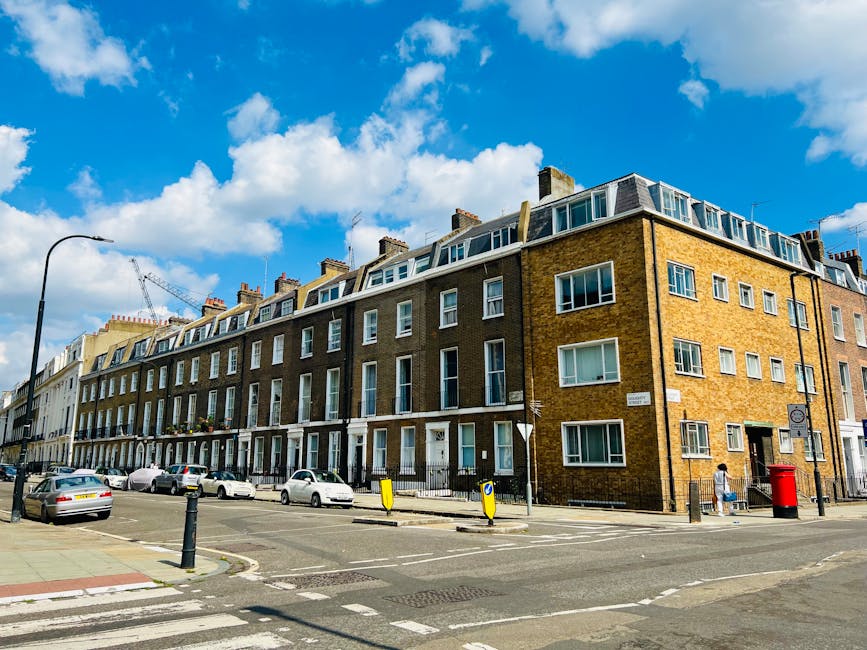 Exterior view of a multi-story residential building in Hampstead under a bright blue sky with scattered clouds. The building features traditional brickwork with large rectangular windows, some with white frames, and a row of small dormer windows on the top floor. The ground level has individual entrance doors, with a sidewalk running along the front. Several parked cars line the street, and a red postbox and street lamps are visible. The scene is well-lit, showcasing a clean and tidy environment, reflecting a typical London street scene. Cleaner Hampstead specializes in domestic cleaning services, including surface cleaning and deep cleaning, ensuring properties stay hygienic and well-maintained in such residential areas.