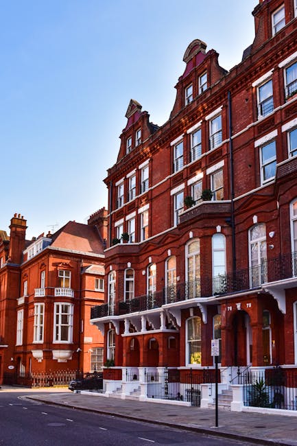 Exterior view of a red brick residential building on Flask Walk NW3 in Hampstead, featuring multiple large sash windows with white frames and decorative arched brickwork. The building has a white awning with ornate decorative brackets above the ground-floor entrance, a small black metal fence enclosing the front steps, and a clear blue sky overhead. The street is quiet with minimal traffic, and neighboring buildings with similar architectural style are visible. No cleaning tools or surfaces are visible in this exterior shot. Cleaner Hampstead specializes in domestic cleaning and property maintenance, ensuring homes like these are kept spotless and well-maintained.