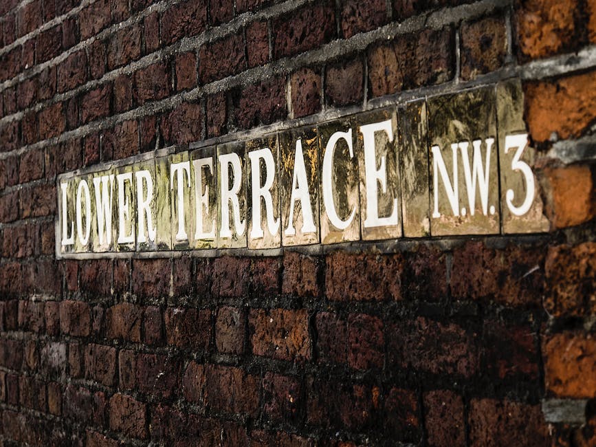A close-up view of a brick exterior wall with a metal street sign attached, reading 'LOWER TERRACE NW3' in white letters on a green background. The bricks are dark red with visible mortar joints, and