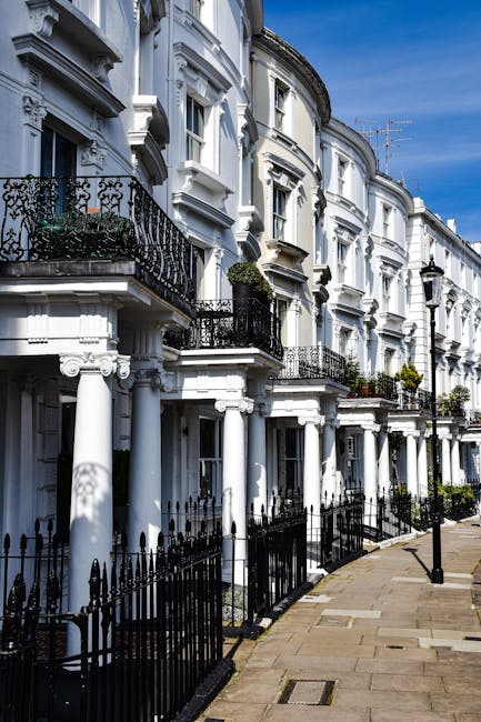 A row of white terraced houses featuring ornate architectural details, including decorative moldings and black wrought iron balconies, situated along a paved street with black metal railings. The surfaces of the buildings appear clean and well-maintained under natural daylight, with clear glass windows reflecting the sky. Potted plants and small floral arrangements are visible on some balconies, contributing to the tidy residential environment. The overall scene showcases the pristine condition of the property exteriors, emphasizing meticulous exterior surface cleaning and maintenance typical of professional domestic and commercial cleaning services offered by Cleaner Hampstead near Hampstead Heath.