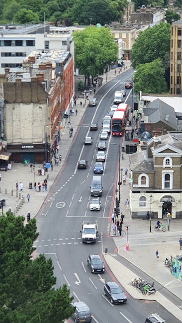 An aerial view of a city street in Hampstead, featuring a mix of historic and modern buildings. The street is lined with lush green trees and has a row of parked cars along the curb. Moving vehicles, including buses and cars, are driving down the road. Pedestrians are walking on the sidewalks, and bicycles are parked near a small building with classic architecture. Bright daylight illuminates the scene, providing clear visibility of the urban environment. The image reflects a typical bustling residential and commercial area in NW3, showcasing well-maintained surfaces with clean streets and organized traffic, consistent with the professional cleaning and maintenance standards offered by Cleaner Hampstead.