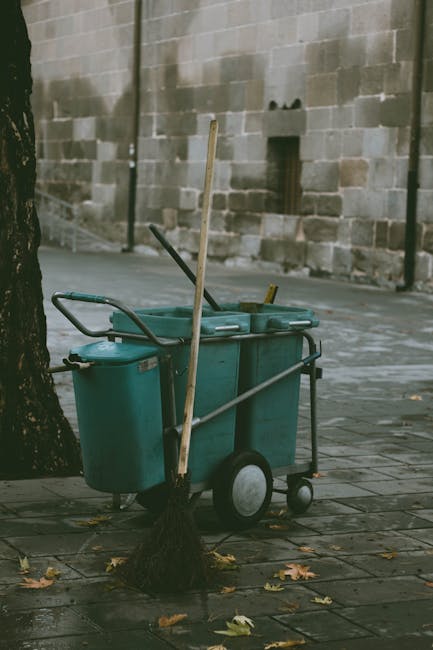 A cleaning trolley parked on a paved outdoor area next to a tree, with a brick wall in the background. The trolley contains various cleaning tools, including a mop with a wooden handle, a duster, and cleaning cloths. The trolley's compartments are made of plastic, and it has large wheels for mobility. The scene shows an urban environment with fallen leaves scattered on the ground, and the lighting suggests daytime. This image exemplifies surface cleaning and maintenance in an outdoor public space, aligning with services offered by Cleaner Hampstead for comprehensive cleaning solutions.